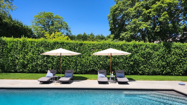 Ein Swimmingpool mit zwei Liegestühlen und Sonnenschirmen, umgeben von einer Hecke und Bäumen.