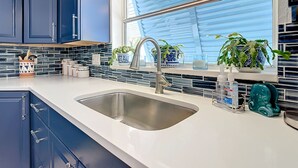 A modern kitchen with dark cabinets, a white countertop, a stainless steel sink, and a window with blinds. - Mermaids' BeachHouse: Walk2Beach • Pool • KingBed (Cocoa Beach)