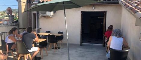 Un patio al aire libre con mesas y sillas, un paraguas verde y un edificio con tejado de baldosas al fondo.