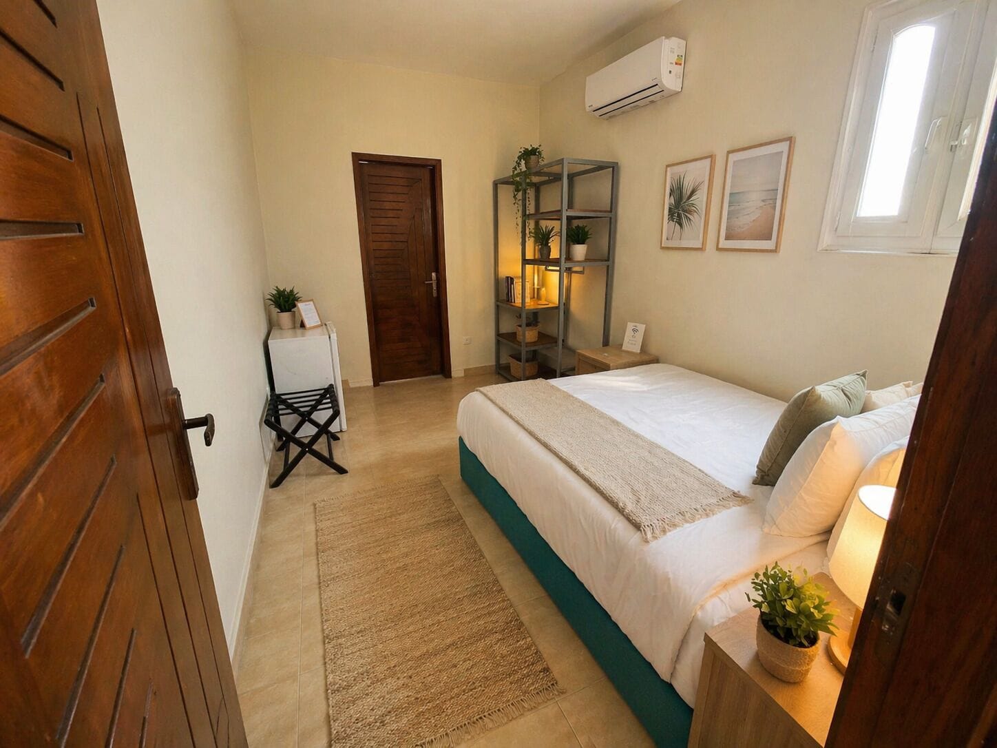 Un dormitorio con cama, una mesita, una estantería con plantas, una puerta y un equipo de aire acondicionado.