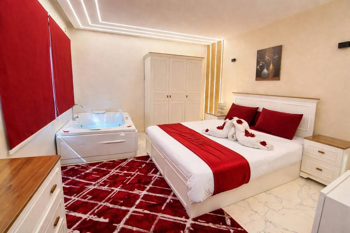 A hotel room with a bed, a red and white patterned rug, a white wardrobe, a white cabinet, and a white bathtub with a shower head.
