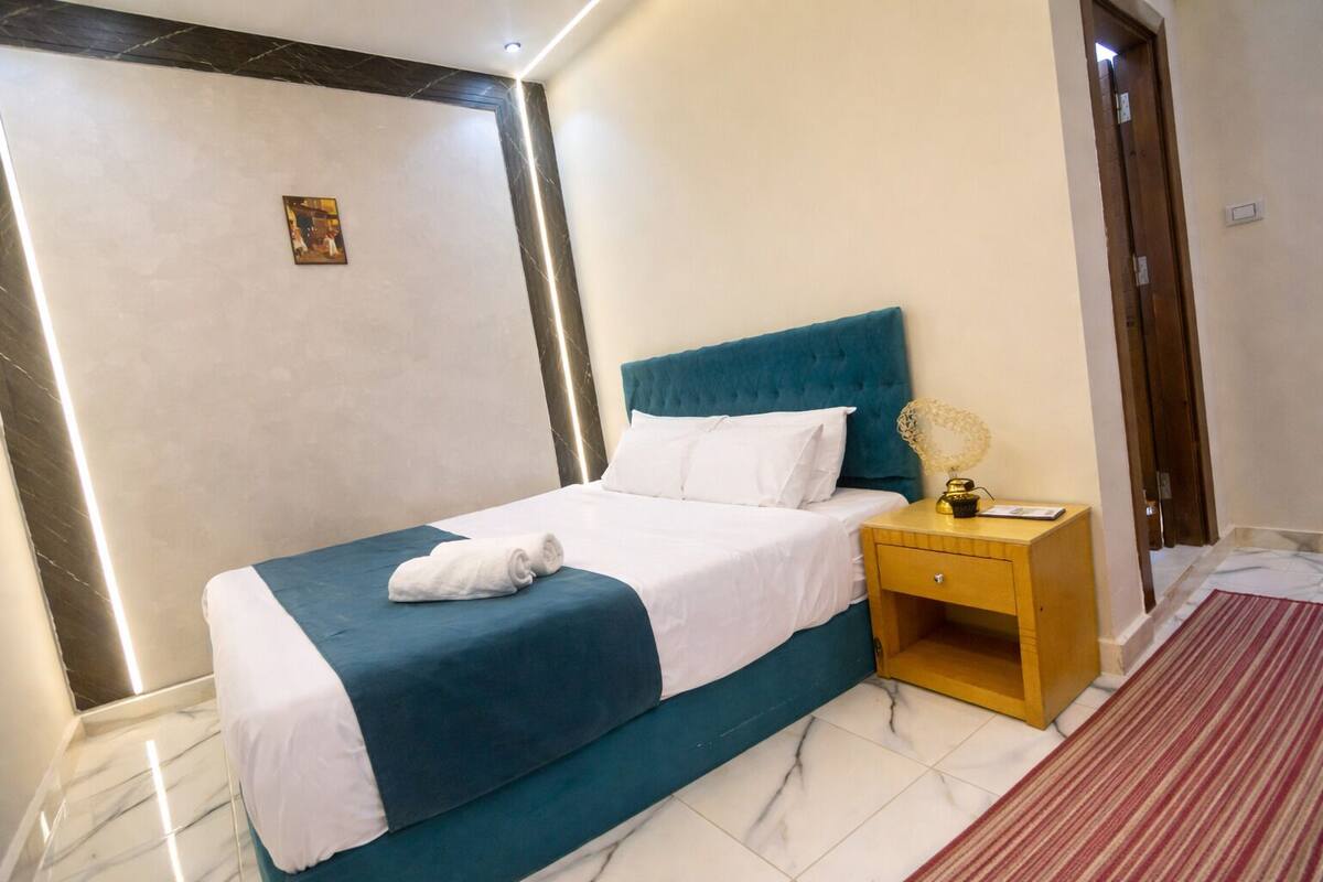 A neatly made bed with a blue headboard, a bedside table with a lamp, and a small picture on the wall.