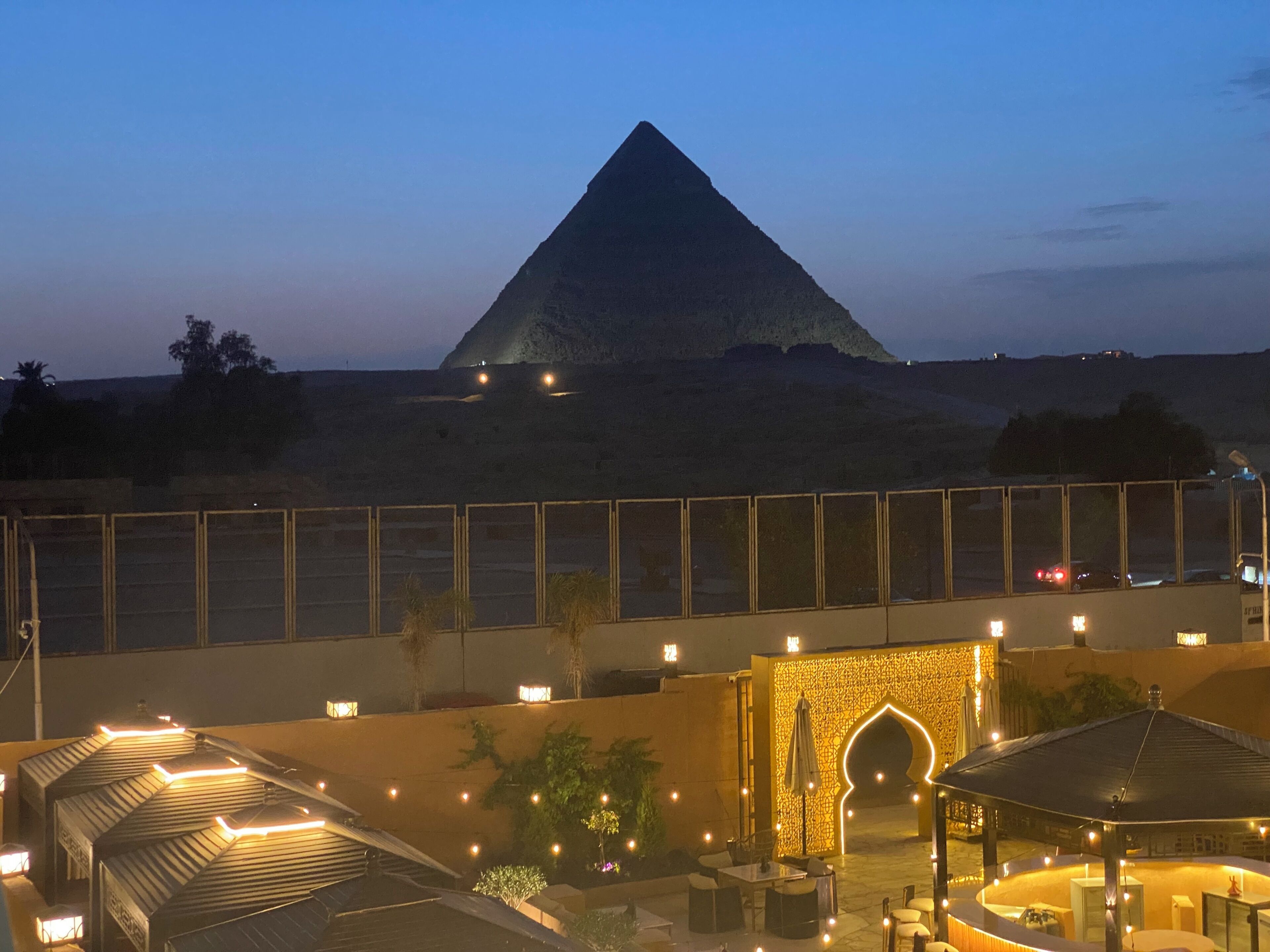 A pyramid with a fence in the foreground, a covered outdoor seating area, and a building with a patterned facade.