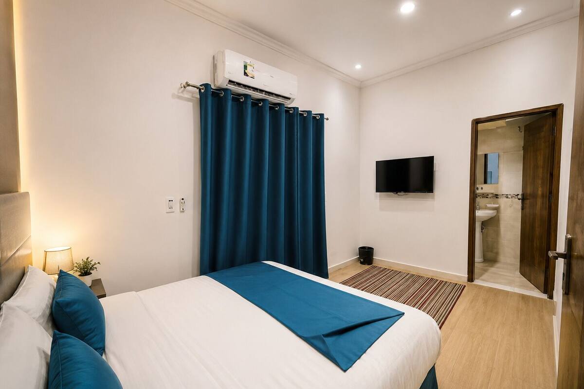 A hotel room with a bed, an air conditioning unit, a television, and a bathroom visible through an open door.