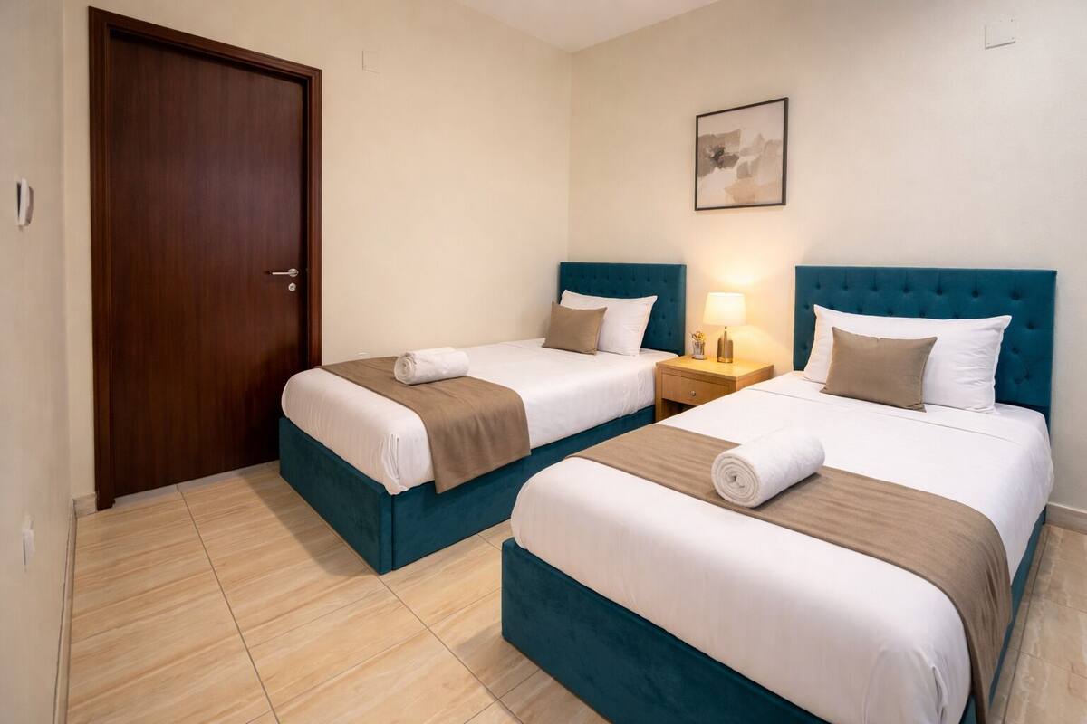 A hotel room with two beds, a wooden door, a nightstand with a lamp, and a framed picture on the wall.