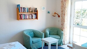 A room with a bed, a small table, two green chairs, a bookshelf, and a window with curtains. - Heckenrose 5 EG Rechts (Norddorf auf Amrum)