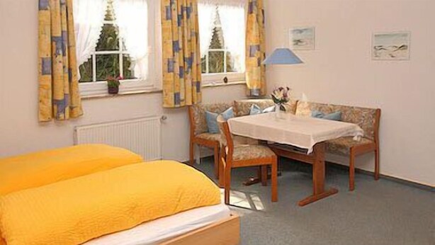 Une pièce comprenant un lit, une table et un banc. On y trouve des fenêtres avec des rideaux, un radiateur et une lampe.