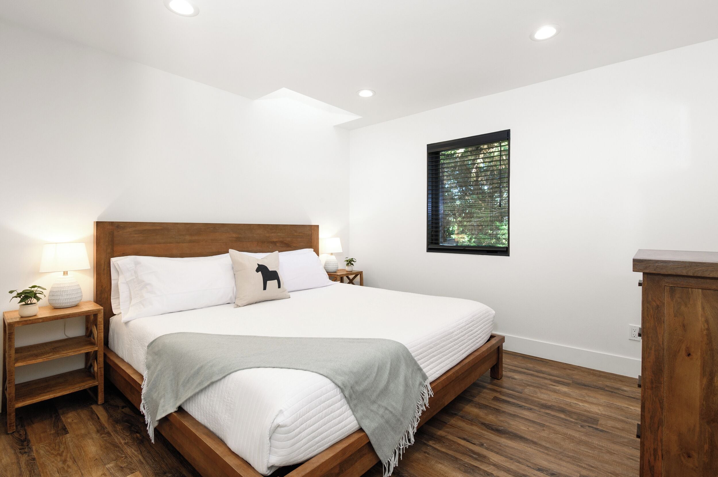 Una camera da letto con una struttura letto in legno, biancheria da letto bianca, un comodino con una lampada, una piccola pianta e una finestra con vista sugli alberi.
