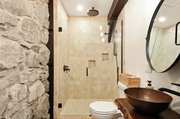 Une salle de bain moderne avec un mur d’accent en pierre, une douche à parois de verre, un lavabo en forme de bol en bois et des toilettes.