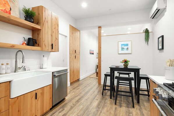 A modern kitchen with wooden cabinets, a stainless steel dishwasher, and a dining area with a black table and stools.