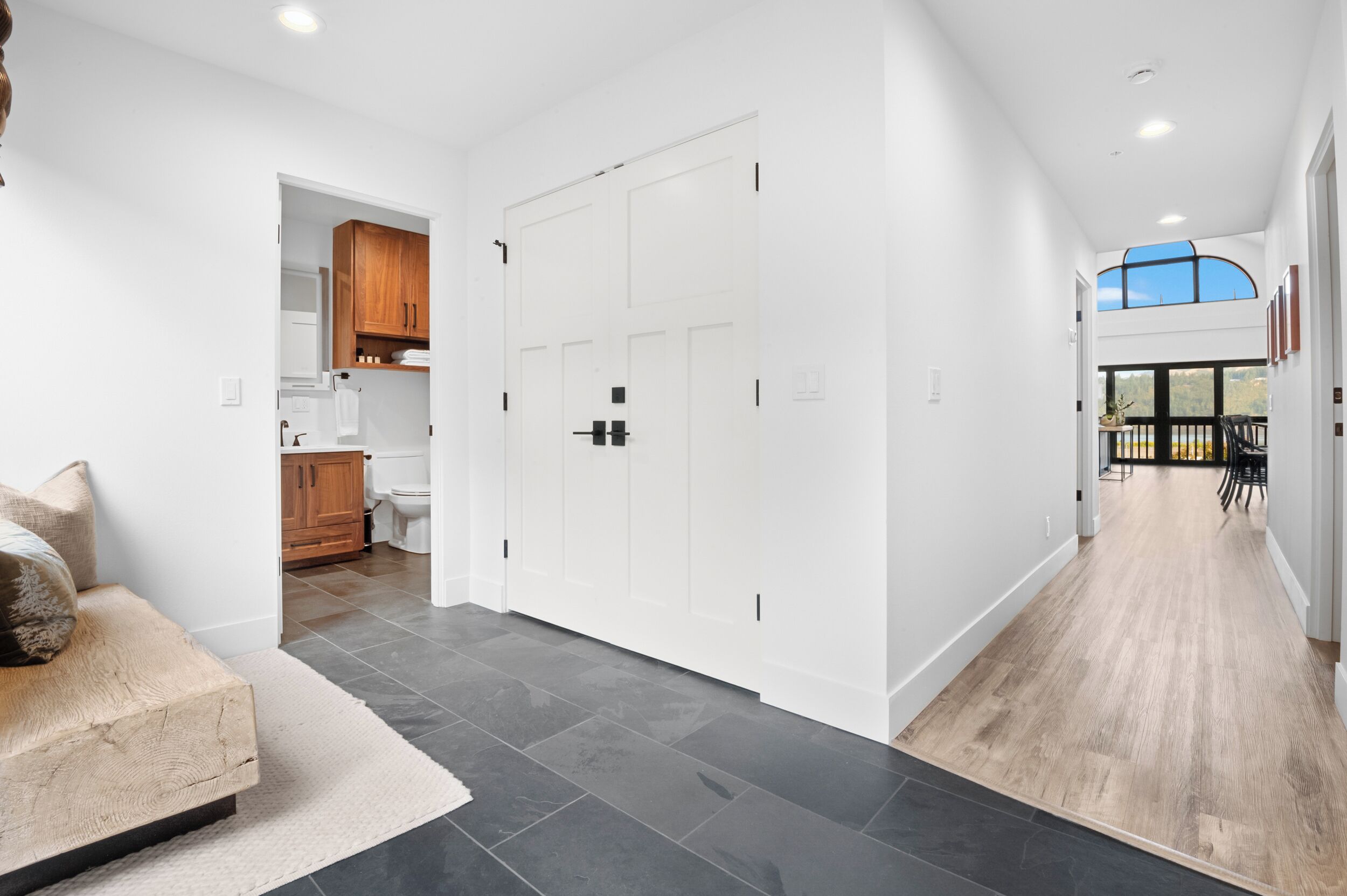 Un intérieur moderne avec un couloir menant à une salle à manger, comprenant une cuisine avec des meubles en bois, une porte blanche et une vue sur l’extérieur.