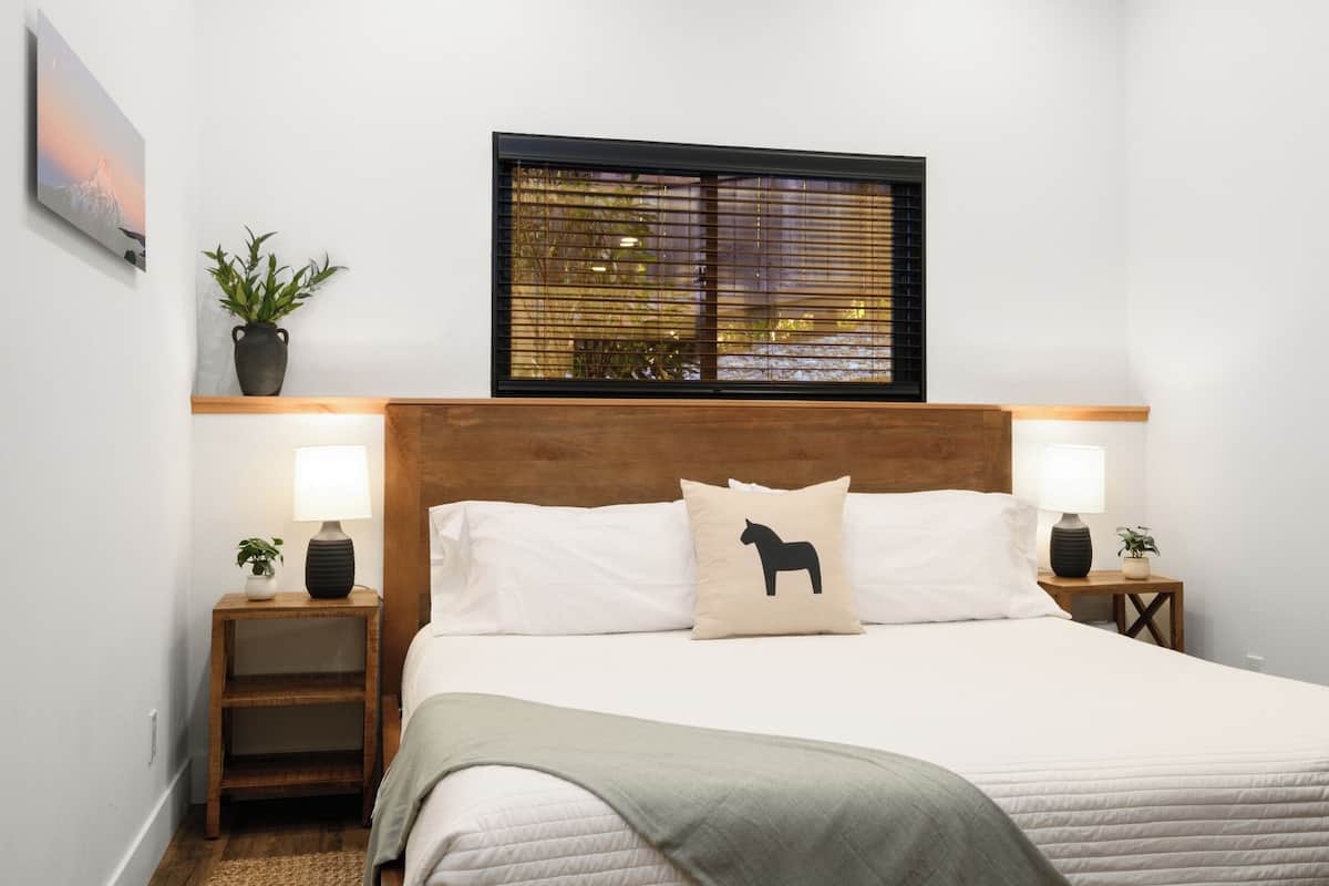 A neatly arranged bedroom with a bed, bedside tables, lamps, a window with blinds, and a wall-mounted picture.