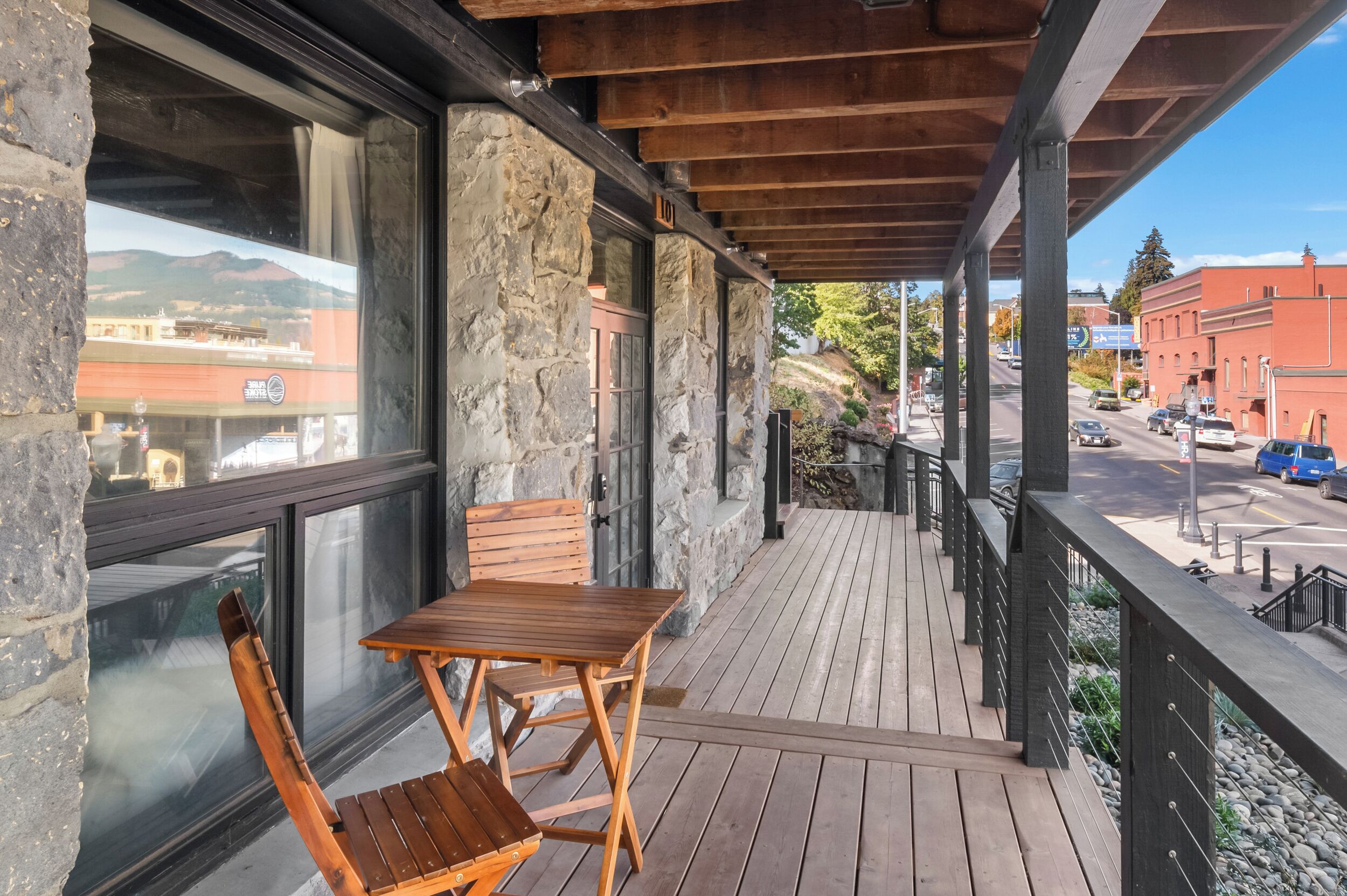 A wooden deck with a table and chairs, stone walls, and a view of a street with cars and buildings.