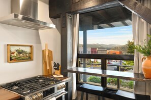 Una cocina con estufa a gas, horno y campana extractora de acero inoxidable. Se observa una ventana con vista a la calle y montañas.