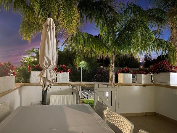 An outdoor patio area with a table and chairs, surrounded by palm trees and flowering plants.