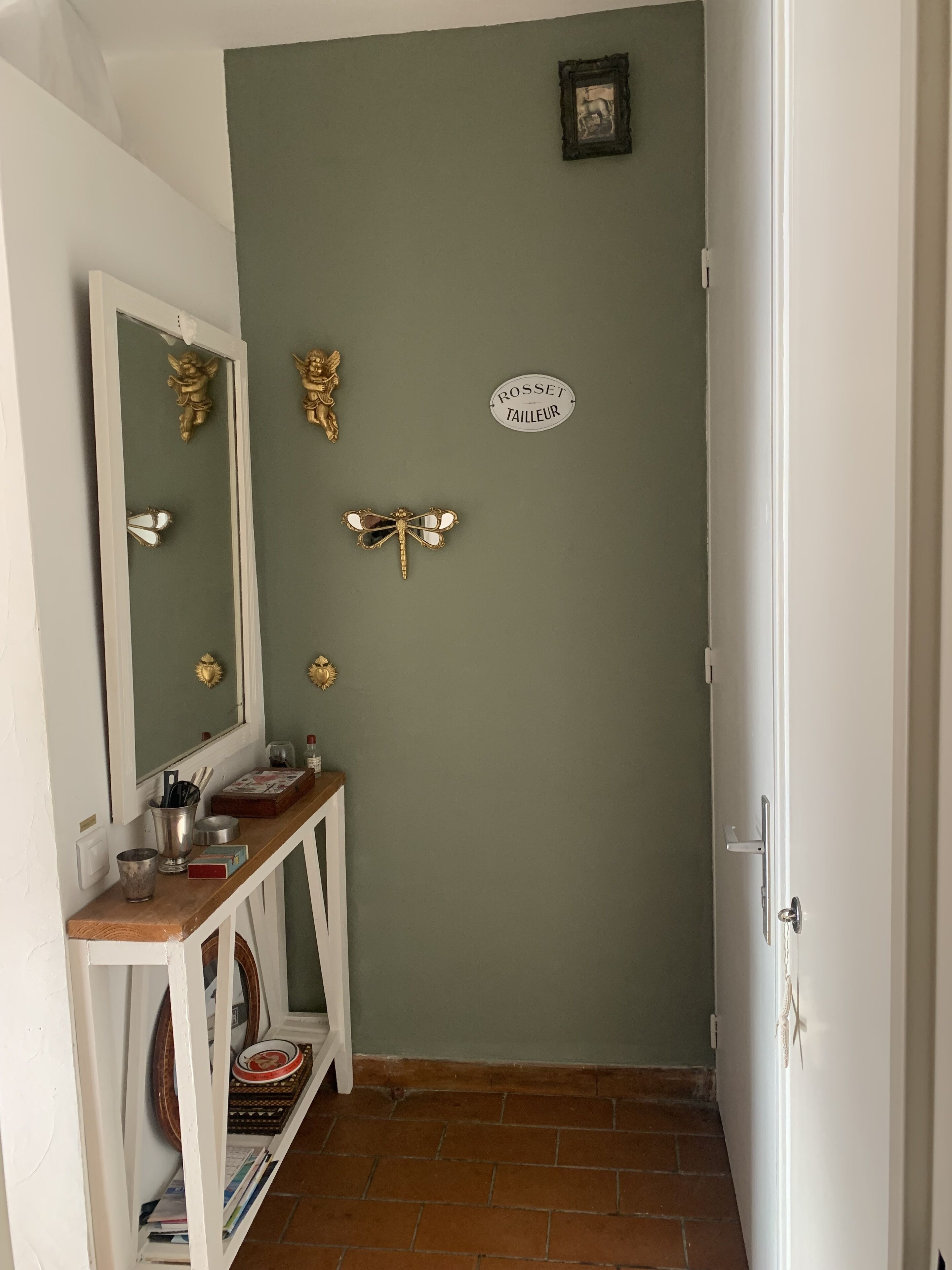 Ein Flur mit grüner Wand, einer weißen Tür und einem Spiegel mit goldenen Engelverzierungen. Ein kleines Regal zeigt verschiedene Gegenstände, und ein Schild trägt die Aufschrift 'Rosset Tailleur'.