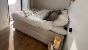 Quarto com cama, um banquinho, luminária na parede e janela com cortinas.