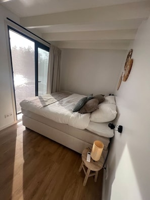 Ett sovrum med en säng, en liten pall, en vägglampa och ett fönster med gardiner.
