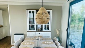 Uma sala de jantar com uma mesa comprida, várias cadeiras, um lustre pendurado e vista para a água através de uma porta de vidro.