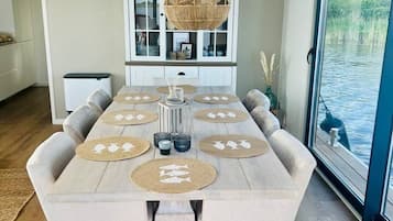 Un espace repas comprenant une longue table, plusieurs chaises, un luminaire suspendu et une vue sur une étendue d’eau, visible à travers une porte vitrée.