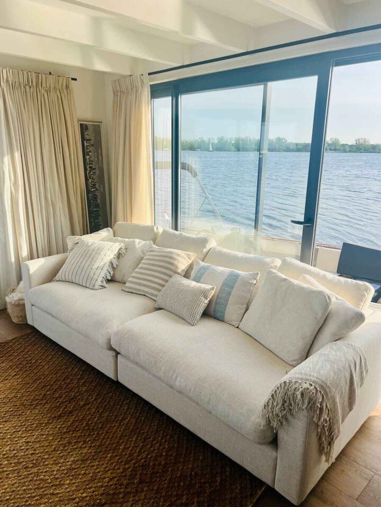 Un salon moderne doté d’une grande fenêtre donnant sur un plan d’eau, d’un canapé blanc agrémenté de plusieurs coussins et d’un tapis à motifs.