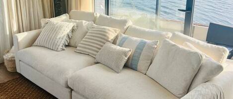 A modern living room with a large window overlooking a body of water, a white sofa with multiple cushions, and a textured rug.
