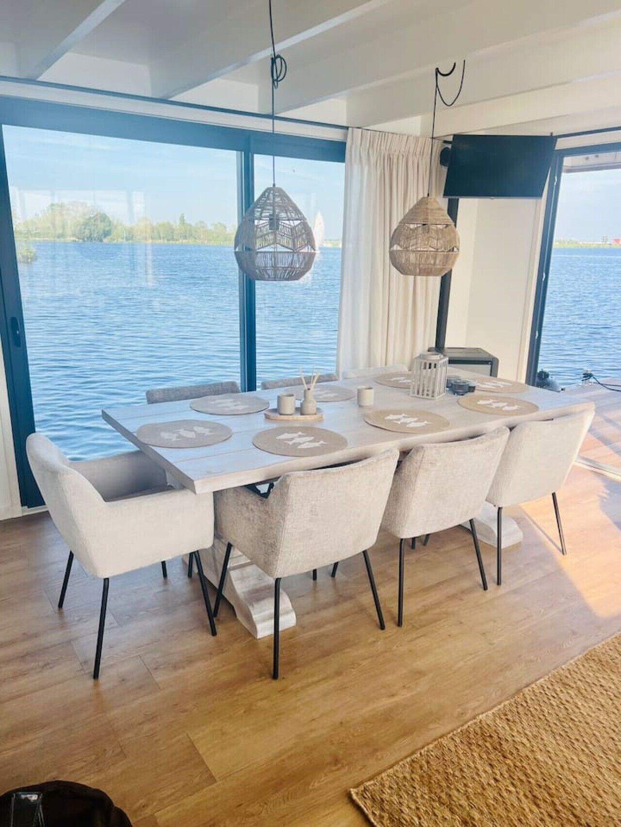 Un espace repas avec une table dressée pour six personnes, de grandes fenêtres offrant une vue sur un plan d’eau et des luminaires suspendus.