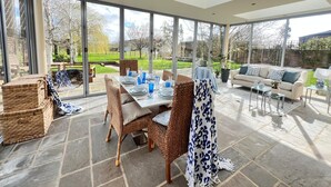 Un amplio patio con mesa de comedor lista para cuatro personas, rodeado de sillas de mimbre y un sofá. Amplias ventanas ofrecen vistas a un jardín y a un lago.