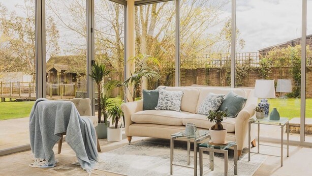 Una sala de estar moderna con un sofá color beige, una mesa de centro de vidrio y vista a un jardín a través de grandes ventanales.