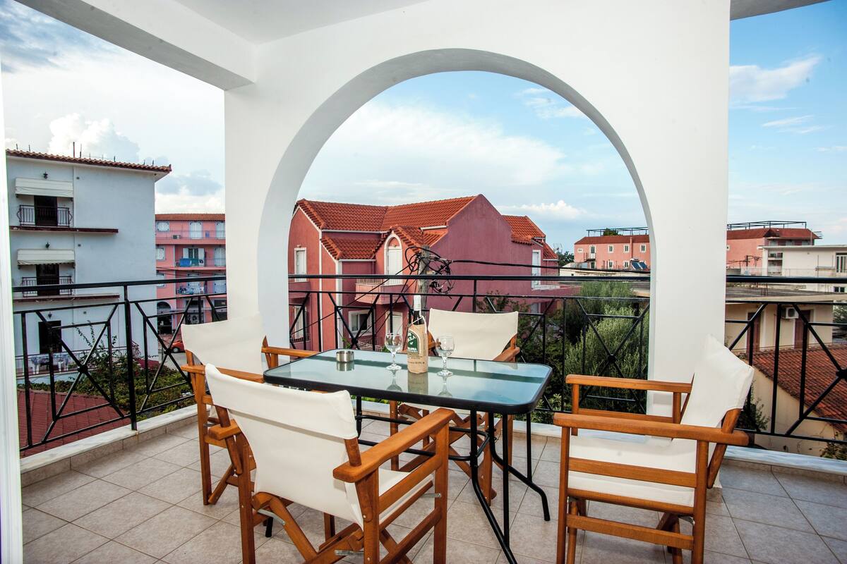A balcony with a glass table and wooden chairs, overlooking a residential area with red-roofed buildings.