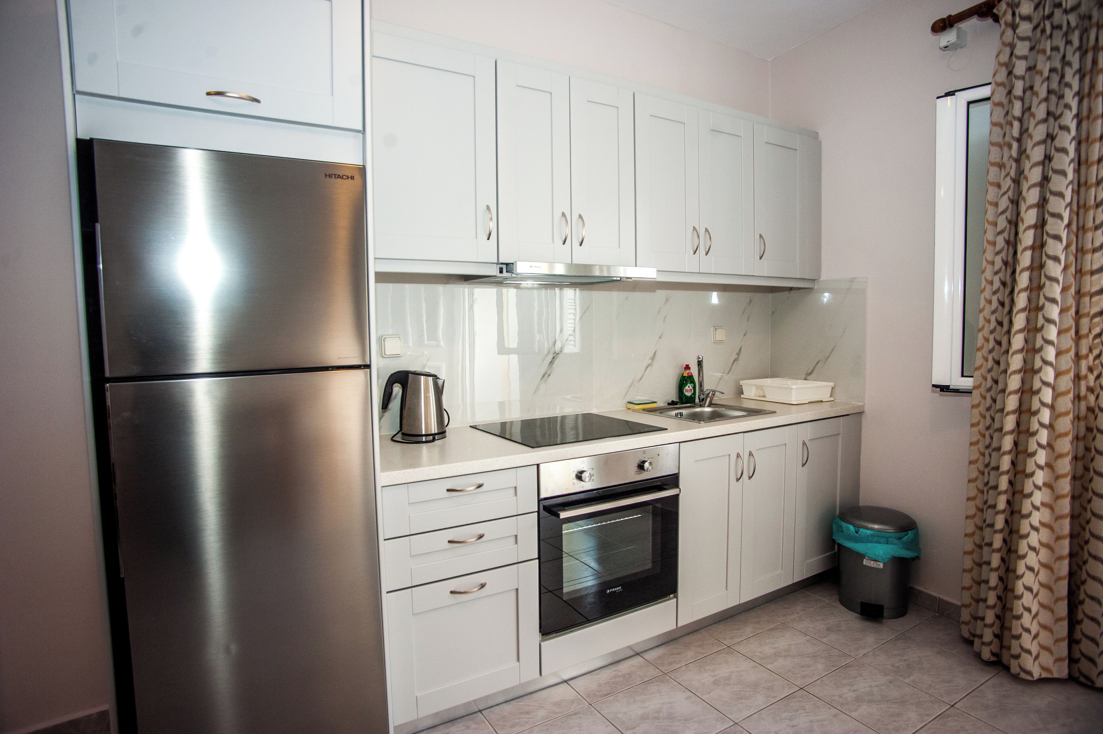 Una cocina compacta con gabinetes blancos, un refrigerador de acero inoxidable, un lavamanos, una estufa y un bote de basura.