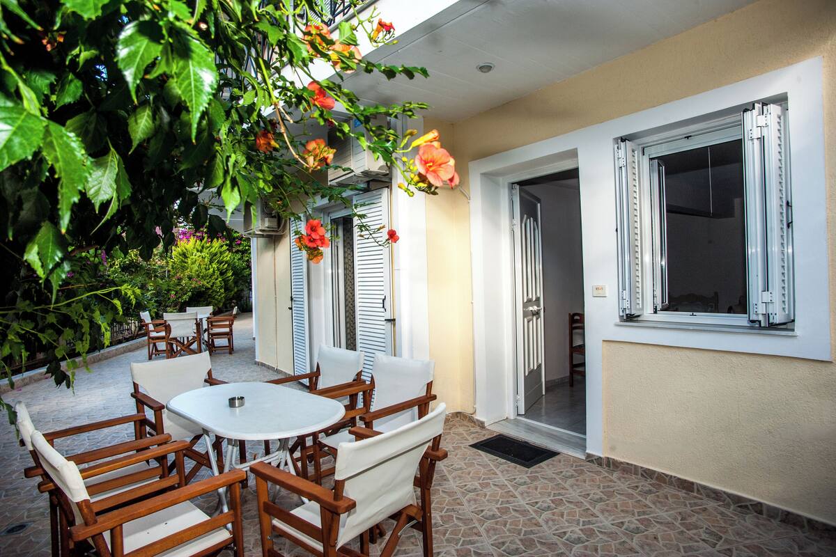 An outdoor patio with wooden furniture and a white table, surrounded by lush greenery and flowers.