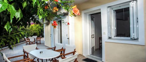 An outdoor patio with wooden furniture and a white table, surrounded by lush greenery and flowers.
