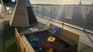 Eine Dachterrasse mit einer Holzspielanlage, einem Sandkasten und einem blauen Eimer.