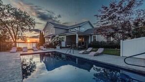 En swimmingpool med liggestole, et hus med en overdækket veranda og en solnedgang i baggrunden.
