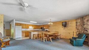 Un vaste espace de vie comprenant une cuisine, des murs en bois, une cheminée en pierre et un ventilateur de plafond.