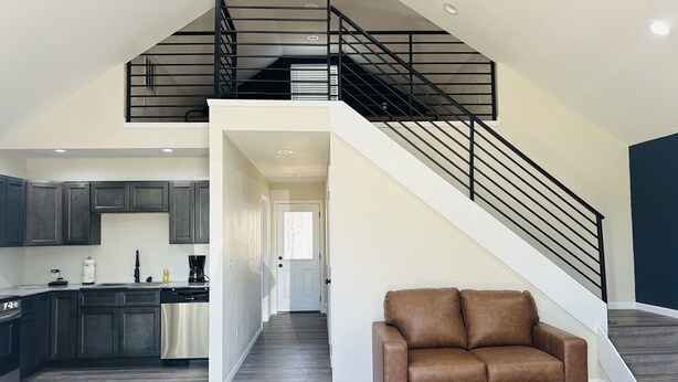 A spacious living area with a brown leather sofa, a kitchen with dark cabinets, and a staircase leading to an upper level. - Loft near Bennett Springs State Park (Lebanon)
