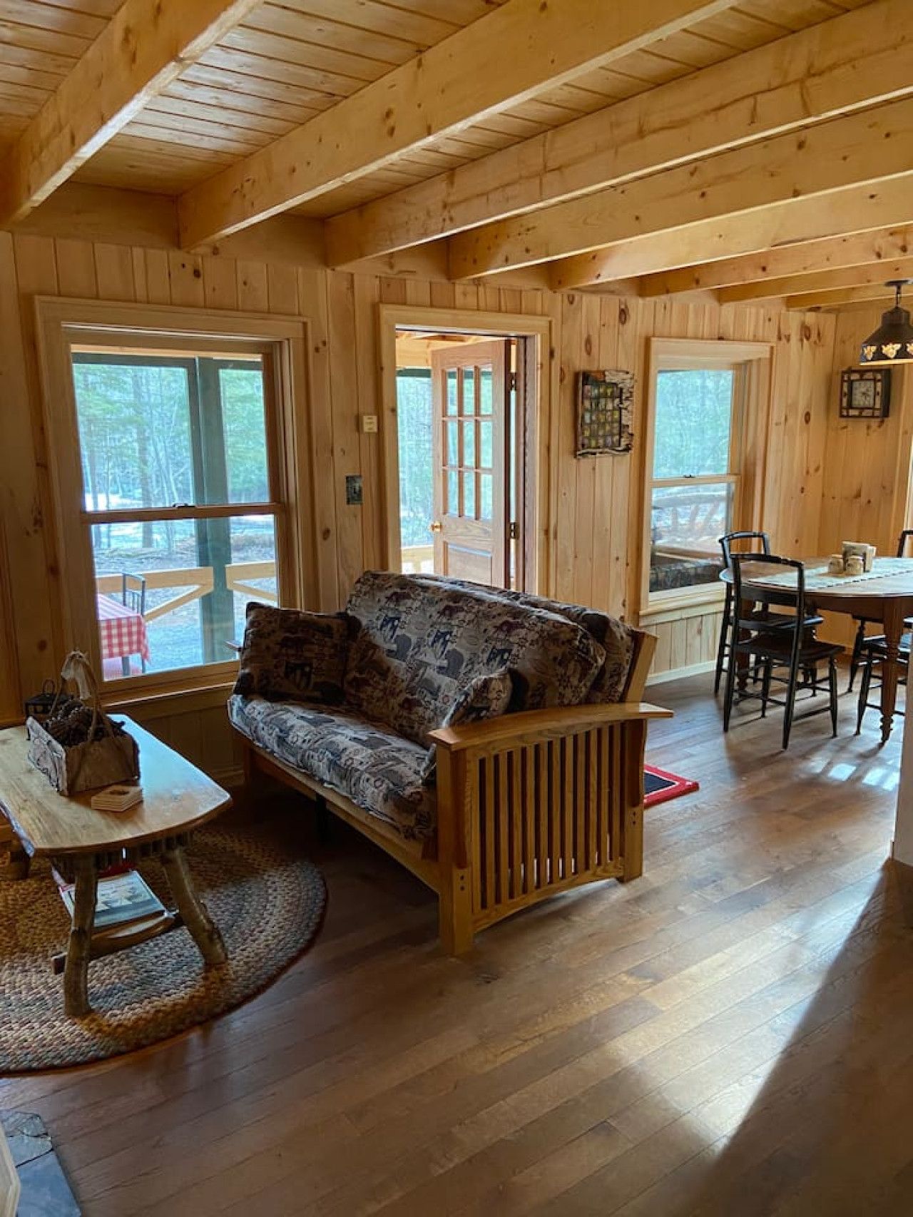 A wooden cabin interior with a sofa, dining table, chairs, and a window overlooking a forest.