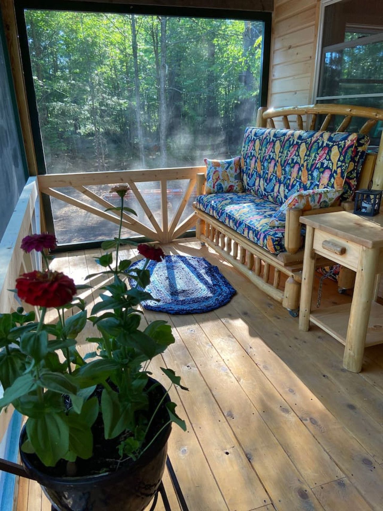 A wooden deck with a sofa and a table, a potted plant, and a view of a forest through a large window.