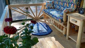Een houten terras met een bank en een tafel, een plant in pot, en uitzicht op een bos door een groot raam.