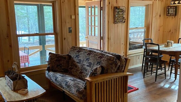 Een houten cabinneerinterieur met een bank, eettafel, stoelen en een raam met uitzicht op een bos.