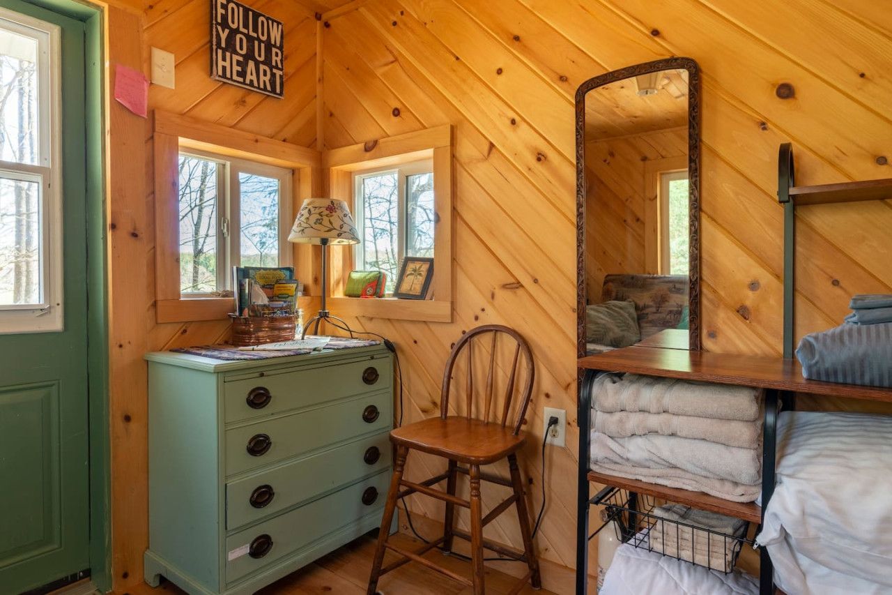 Un interior de cabaña acogedor con techo de madera, un aparador con cajones, una silla, un espejo y una cama nido con mantas.