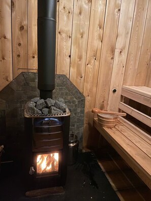 A wood-burning stove with a stone chimney, a bench, and a rustic wooden interior. - The Rock House up in the trees! (Potsdam)