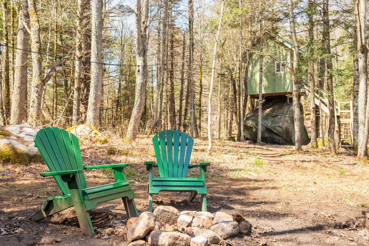 Two green Adirondack chairs in a forested area with a small cabin in the background.