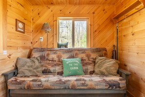 Interior de cabana com sofá estampado com tema de vida selvagem, almofada com a inscrição 'Bem-vindo à Cabana', luminária e janela com vista para uma floresta.