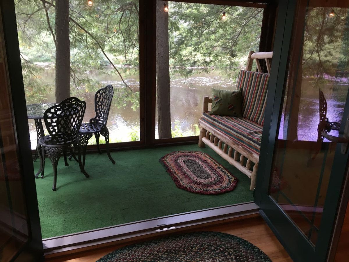 Una habitación con vista a un lago, alfombra verde, banca de madera y sillas de metal.