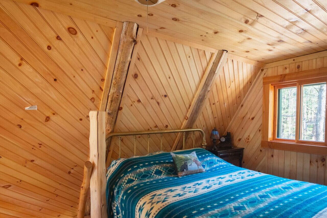 Ein Zimmer einer Holzhütte mit einem Bett, einem Fenster und einer geneigten Decke.
