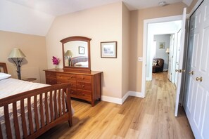 A bedroom with a wooden bed, dresser, and mirror. There is a hallway leading to another room with a chair and lamp.