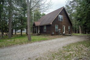 A wooden cabin with a sloped roof, surrounded by trees, and a gravel driveway.