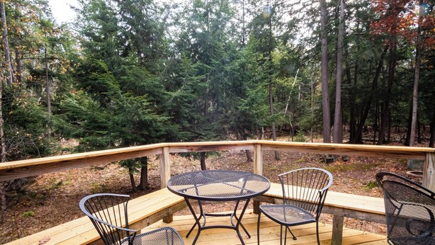 Una terraza de madera con sillas y una mesa de metal, rodeada de un bosque.
